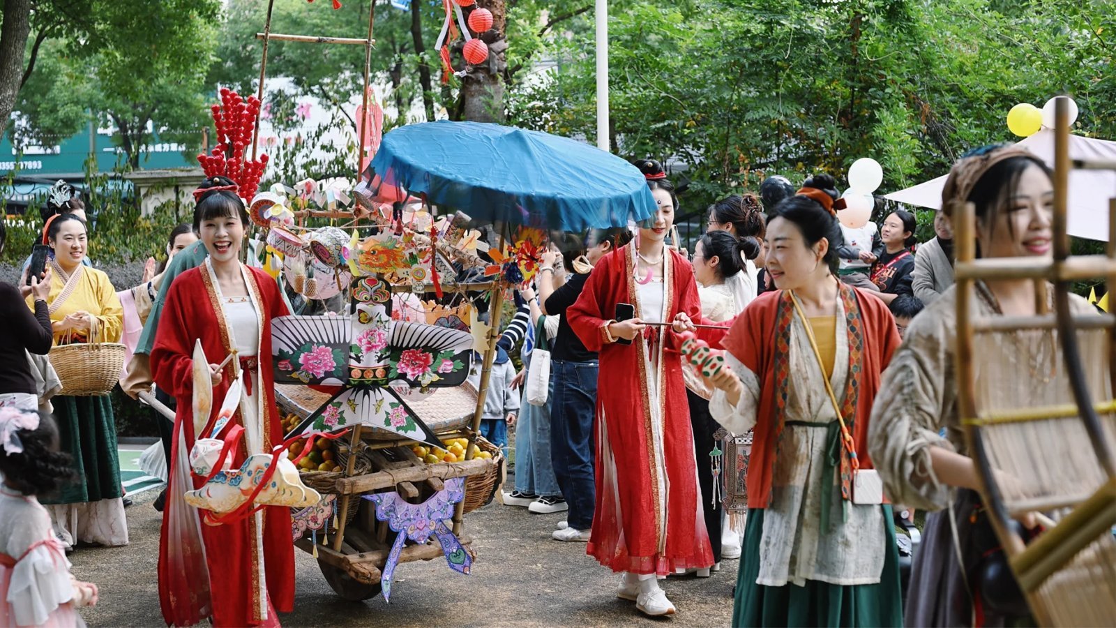 A lively Chinese garden fete — guests in bright hanfu gathered around a wooden cart piled with painted lanterns, paper crafts and pinwheels under a blue paper parasol, red lanterns strung through the green trees — a Chinese festival street event theme