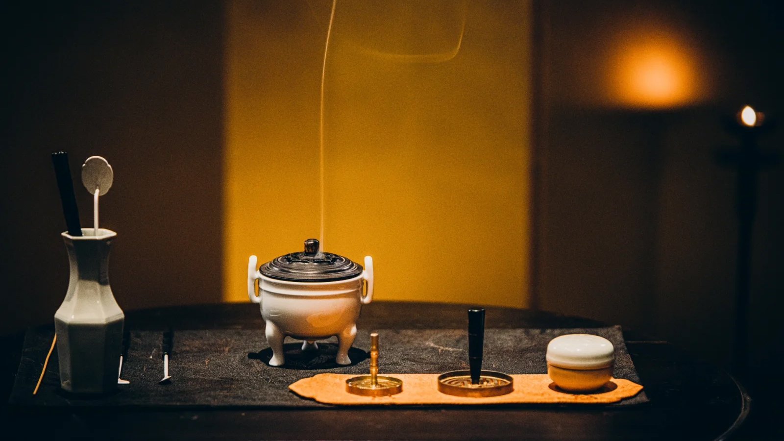 A thread of smoke rising from a white three-legged censer against a dark backdrop, incense tools standing in a celadon vase, a seal mould and paste box laid out on a cloth — a Chinese incense seal workshop theme