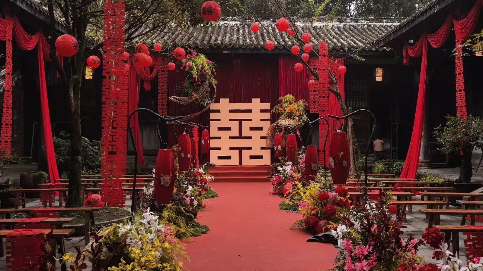 A Chinese wedding courtyard dressed in red silk — a large double-happiness backdrop at the doorway, palace lanterns strung overhead, long tables below in true red and warm gold, and a red wishing tree in the corner