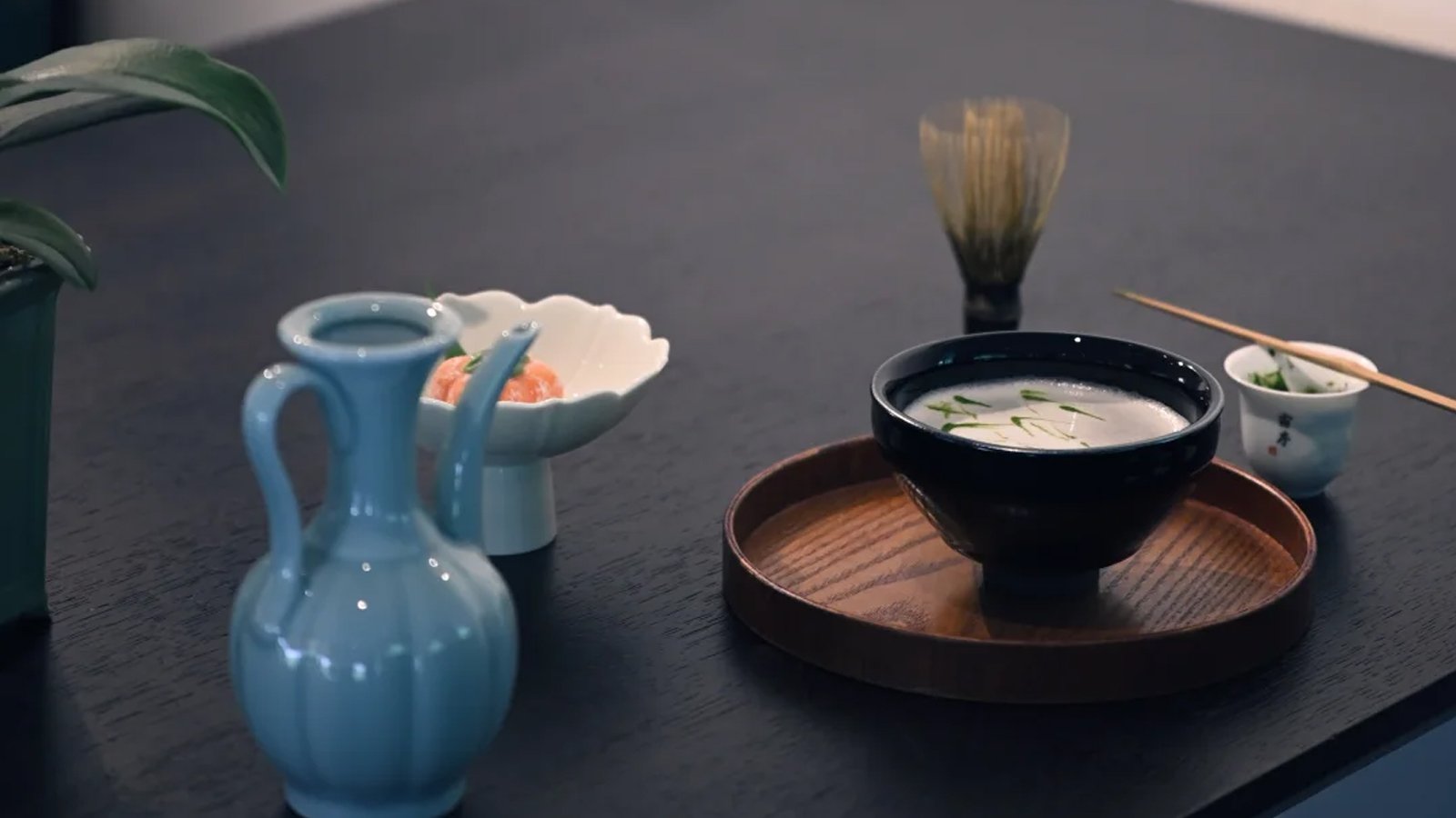A dark-glazed bowl of whisked powdered tea on a wooden tray, with a celadon ewer, a bamboo whisk standing on its stand, a small dish of tea sweets and a spoon on a dark table — a Chinese whisked tea ceremony workshop theme
